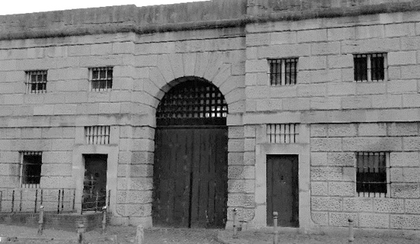 Black and white photo of a grim looking building facade in pale stone with barred windows and massive arched doorway.