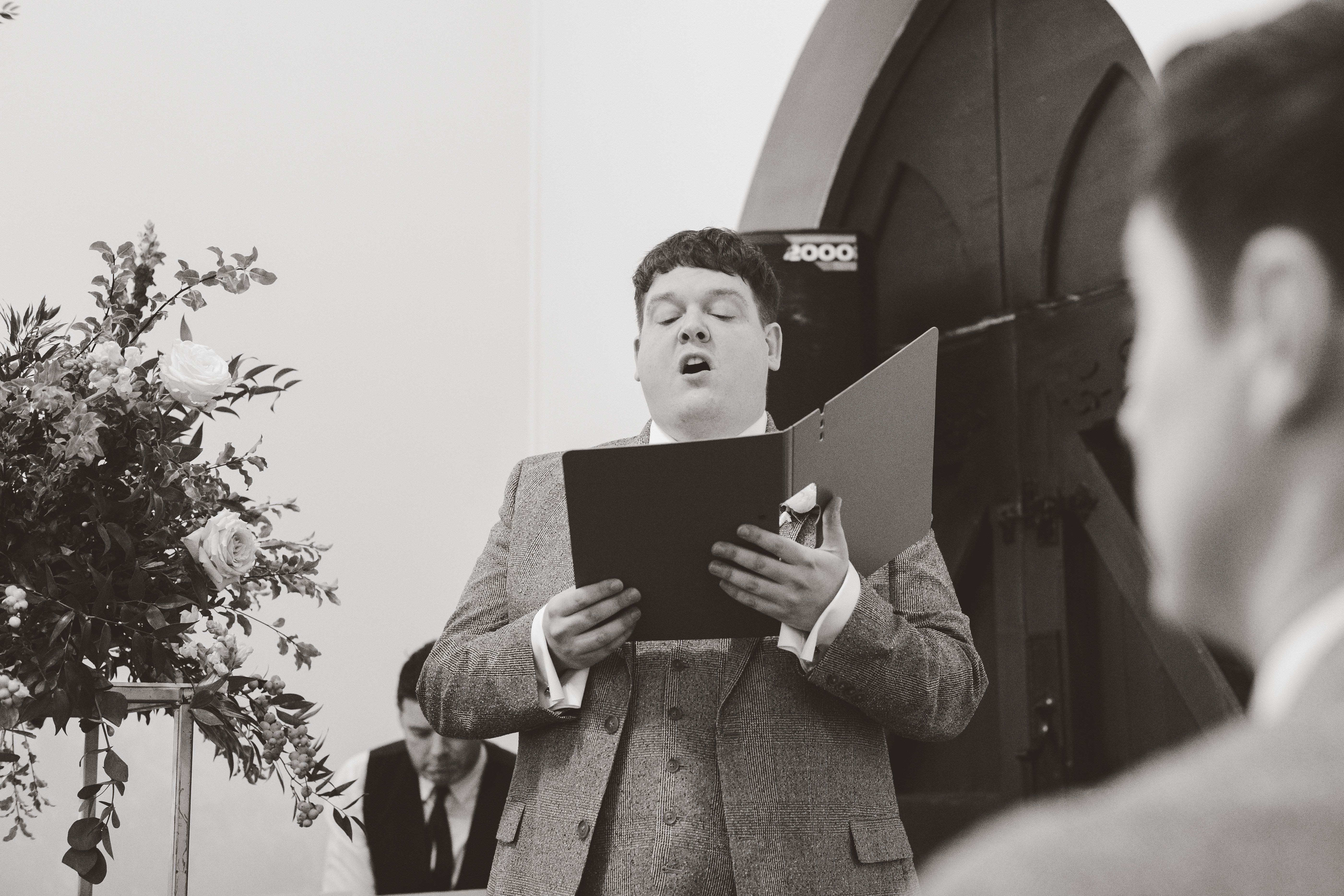 Picture of Chris in a suit with holding a choir sheet and singing at a wedding.