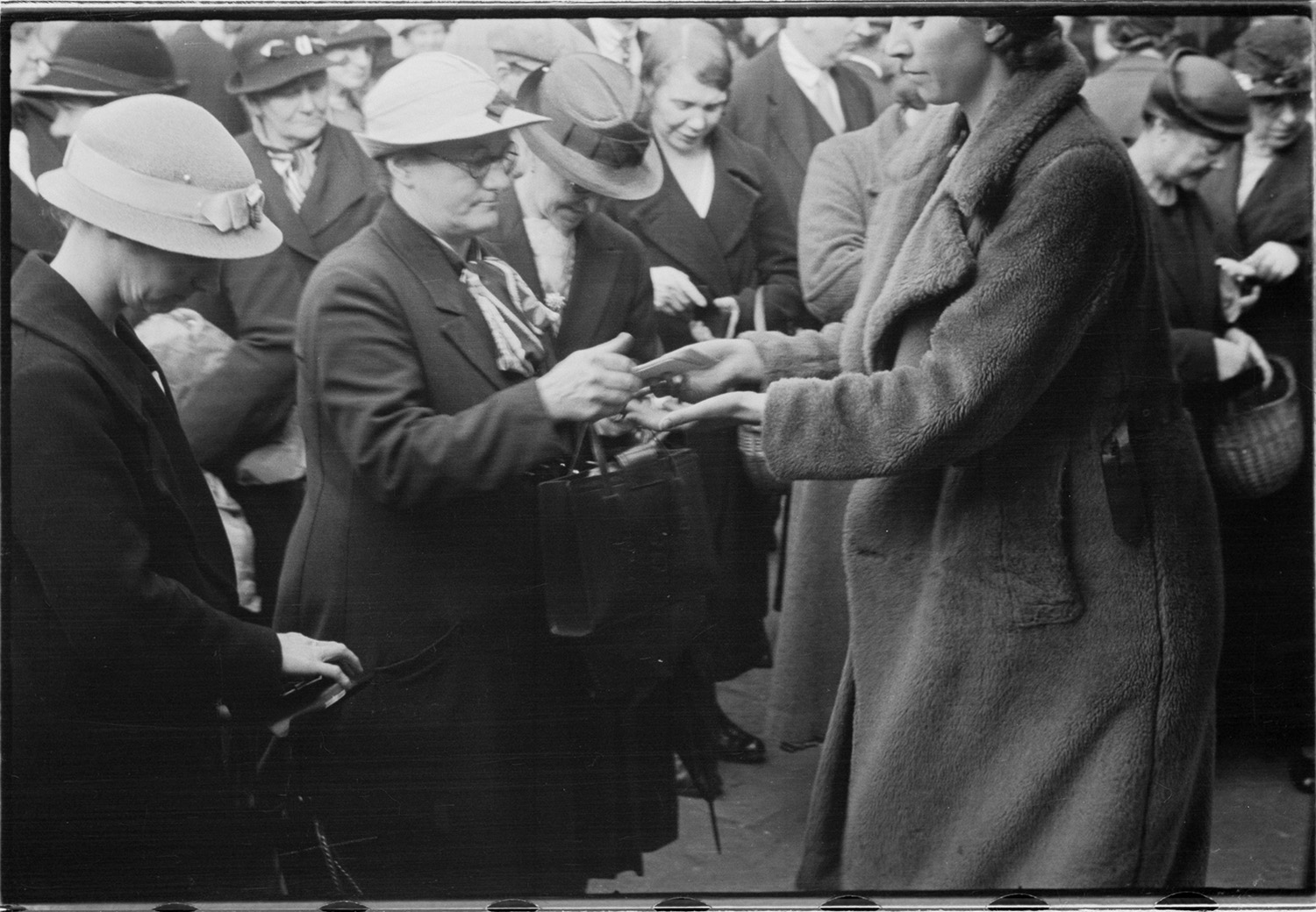 A crowd of mostly middle aged women in coats and hats. In the center a younger woman is exchanging something with one of the women.