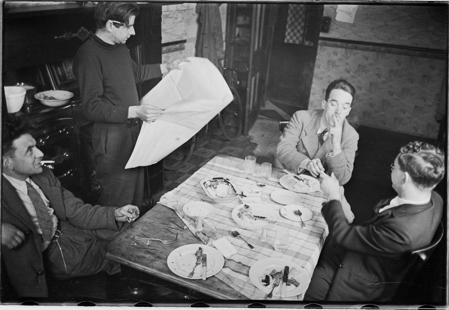 Black and white informal photograph of four middle aged white men sitting around a table of empty plates. One is lighting up a cigarette.