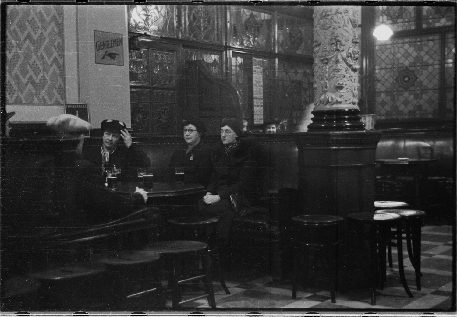 Black and white photo of the interior of an elaborate, old fashioned pub with five people sitting in a booth, three are middle aged women in hats and dark coats. There are 5 pints on the table.
