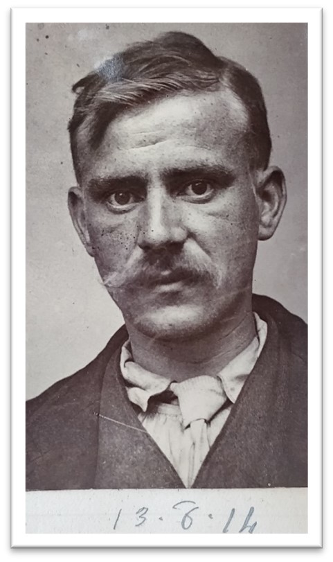 Black and white portrait of a serious looking white man in perhaps his early thirties with short hair in a side parting and a big moustache, he wears a dark sit with a rumpled white shirt and tie