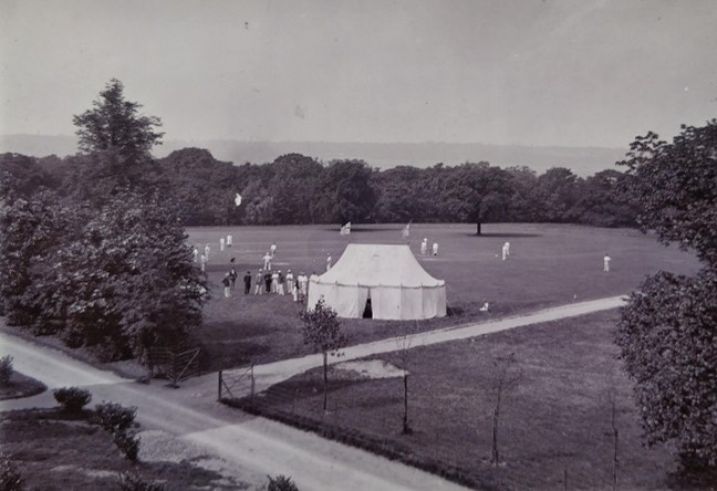 black and white photo of an open space with a white pavilion and cricketers in white scattered over the field