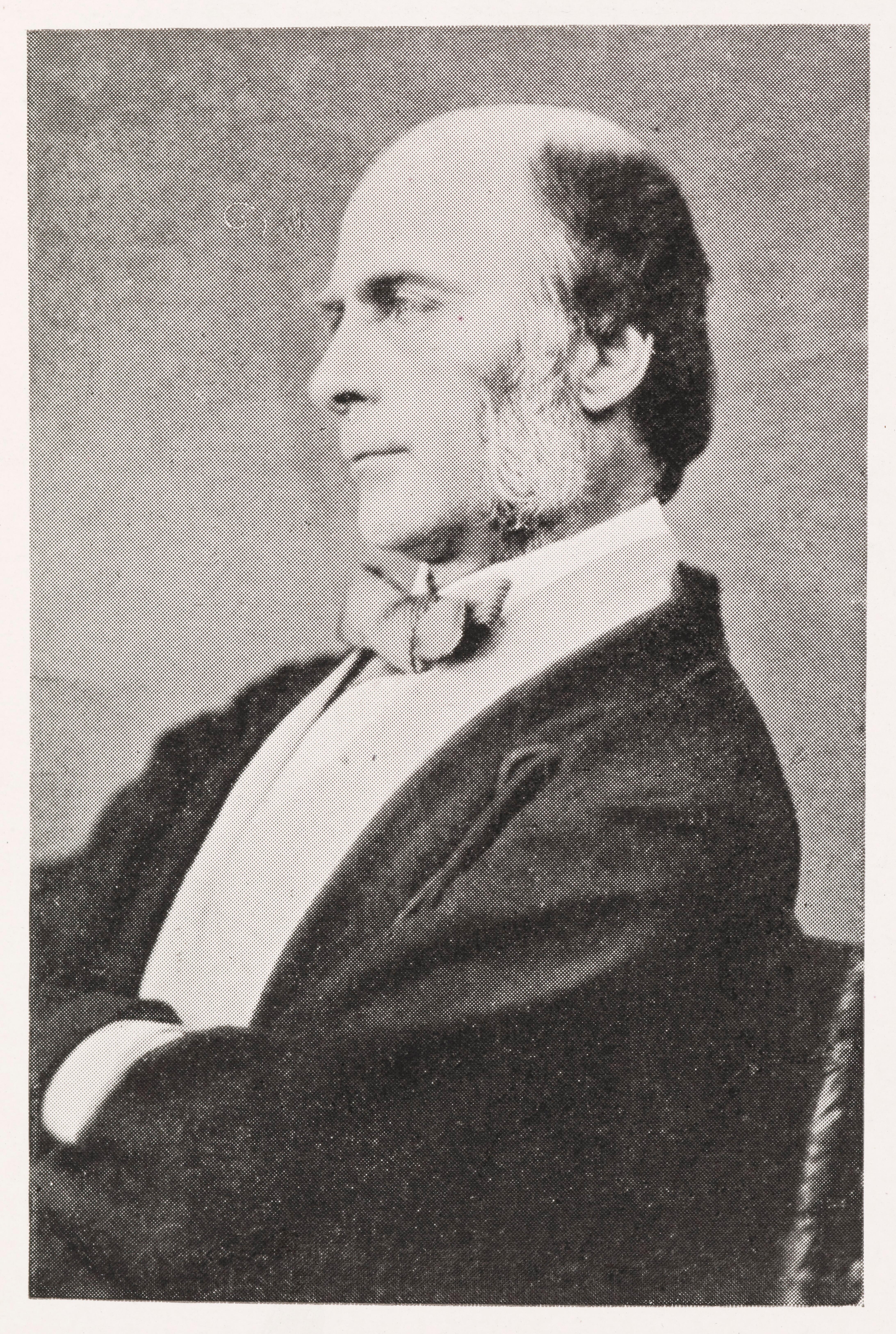 Black and white photo portrait of Francis Galton, shows a white man in profile from the waist up in a bow tie. He has black hair, is bald on the top of his head and white mutton chops.