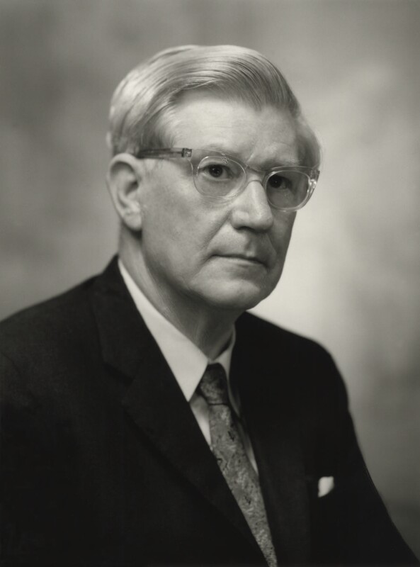 Black and white photo of a white man with white hair and glasses wearing a suit.