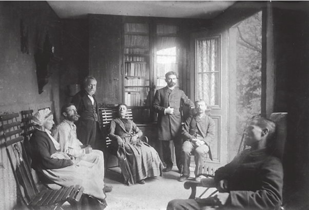 Black and white photo of a small room with 7 people dressed in victorian clothes 5 of whom are seated