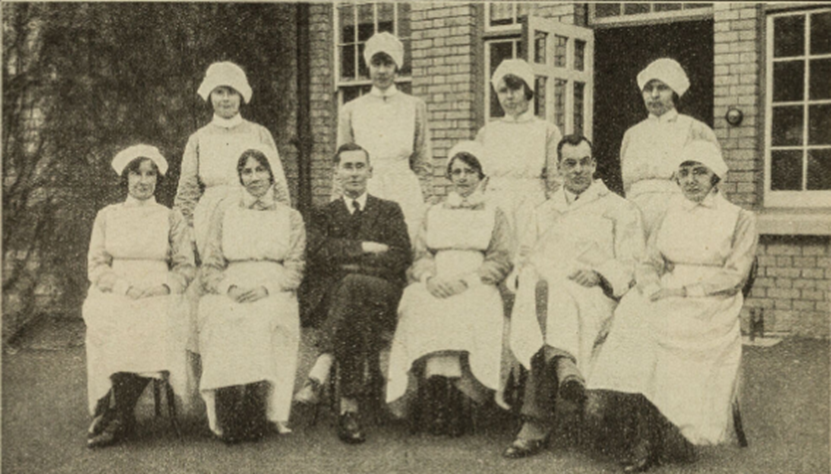 Black and white formal photo of 8 female nurses in old fashioned uniforms and 2 men, one in a suit and one in a white coat. 