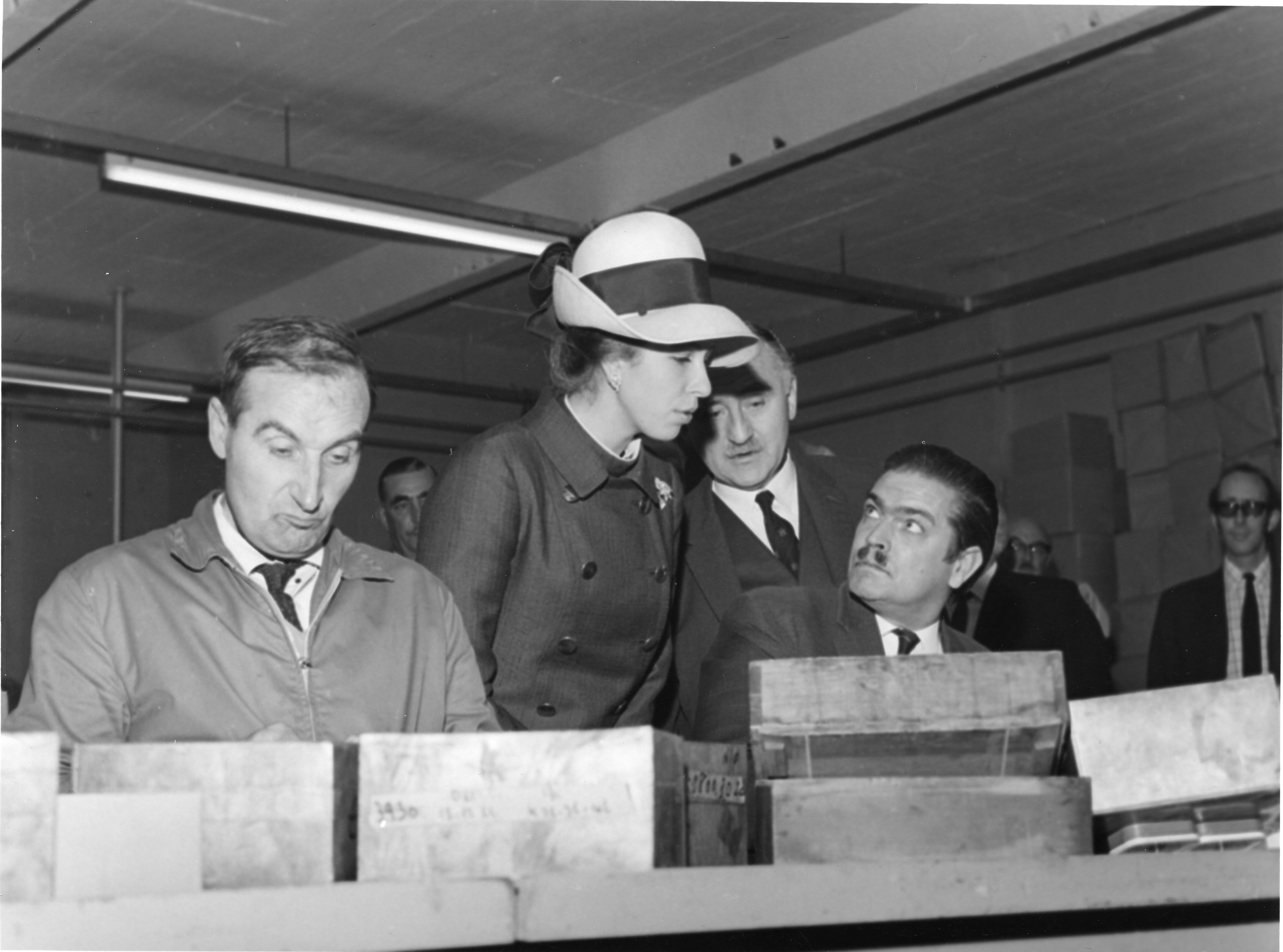 Black and white photo of a young woman in a hat looking over the shoulders of two men with other men in a small crowd behind her. 