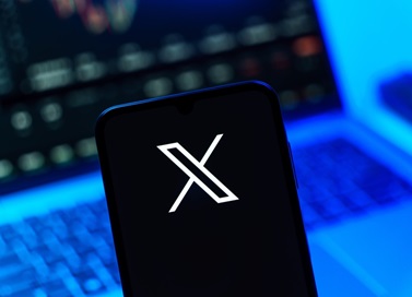X social media logo being shown on a mobile phone with a laptop computer being shown out of focus behind it